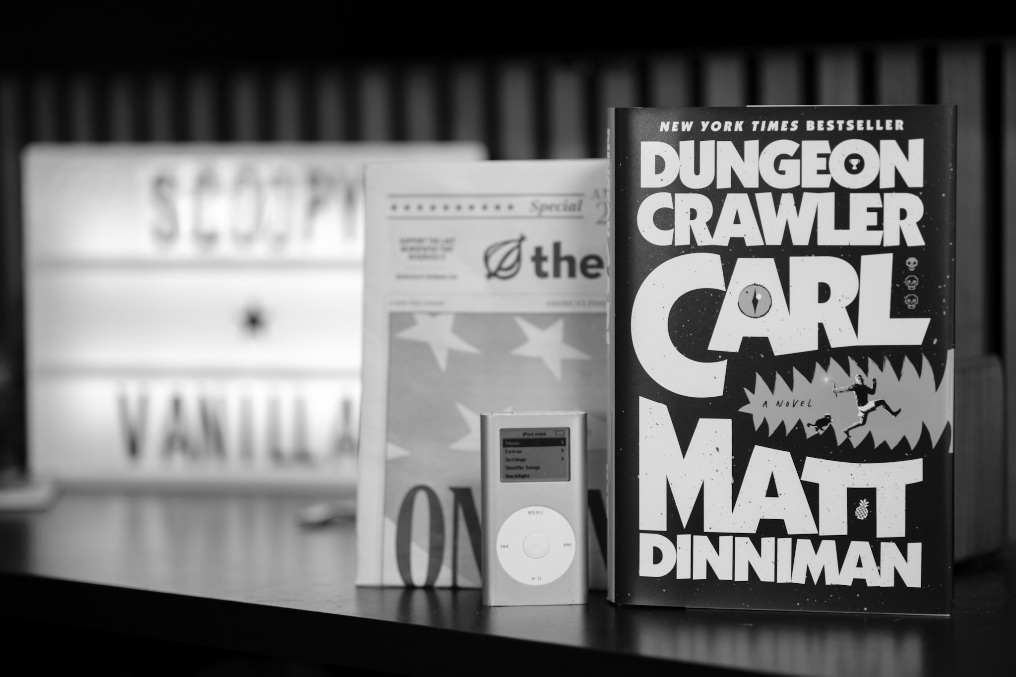Black and white image showing a hardback copy of the book Dungeon Crawler Carl, an iPod Mini, and a newspaper version of The Onion.